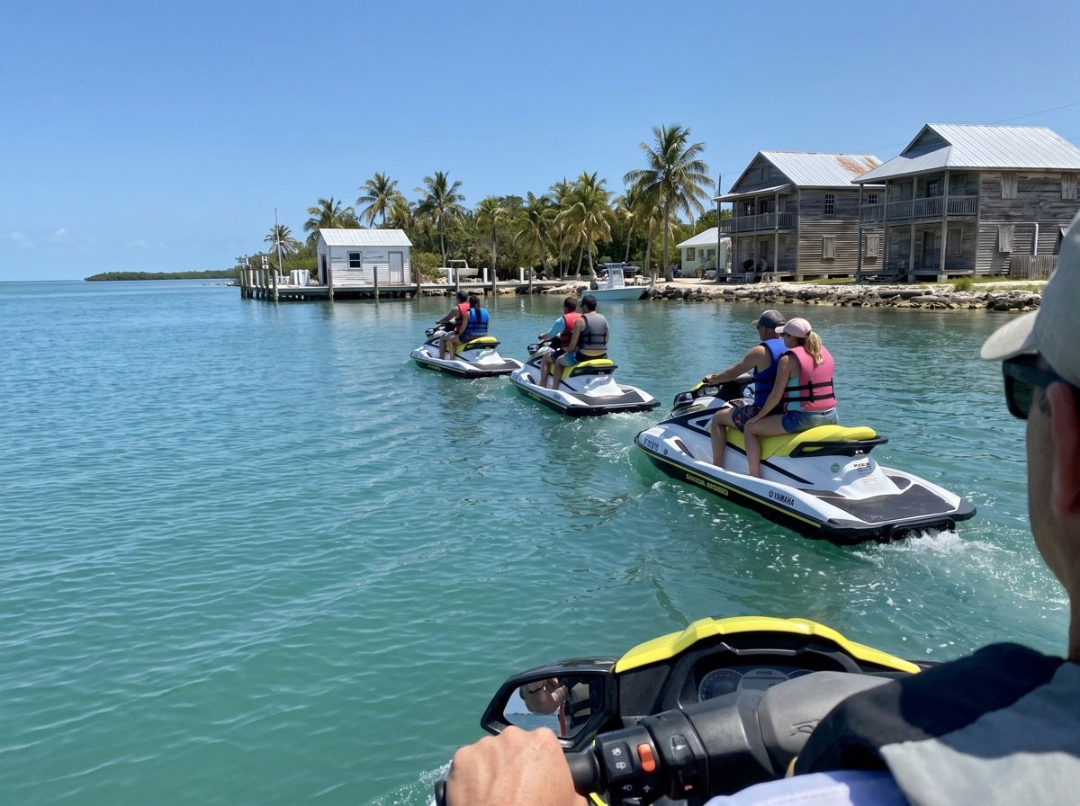 A guide leading a line of jet skis past wooden tin-roof houses on the Keys shoreline